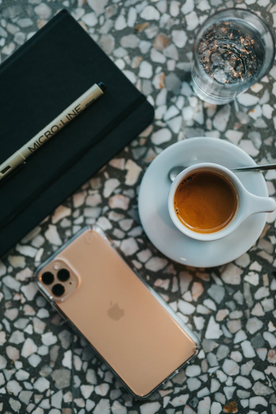Notebook, pen, espresso, water, and phone on terrazzo table.